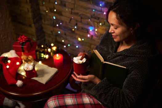 Top view of a happy woman with cup of hot chocolate with marshmallows, enjoying reading book by candlelight, sitting at table with Christmas gifts and sparkling garlands. Magical winter atmosphere