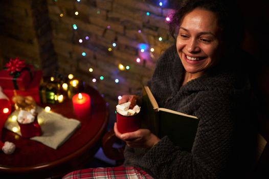 Happy multi-ethnic beautiful woman with cup of hot cocoa with marshmallows, holding a book and smiling a cheerful toothy smile, enjoying magic atmosphere at home during Christmas holidays. December