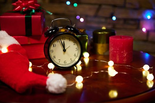 Alarm clock with midnight on the dial, lit garlands, candles and gifts wrapped in decorative shiny wrapping paper, on a wooden table against a brick wall. Magical Christmas and New Year's atmosphere