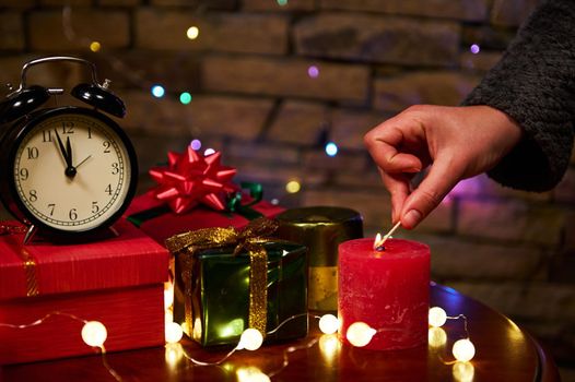 Still life with black alarm clock with midnight on face clock, Christmas presents wrapped in shiny wrapping paper and tied with decorative ribbons, woman's hand lighting festive candle with matches