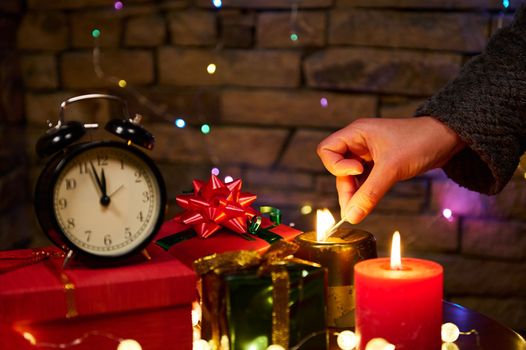 Cozy Christmas background with gifts, and glowing garlands hanging on brick wall. Close-up woman's hand lighting festive candles with matches. New Year's event. Magical atmosphere at winter holidays