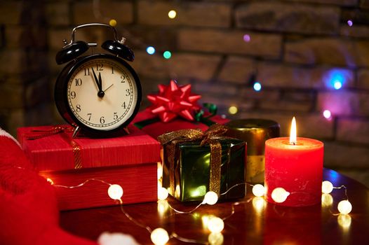Still life with black alarm clock with midnight on dial, Christmas presents wrapped in shiny wrapping paper and tied with decorative ribbons, glowing garland lights on a table. New Year's event. Xmas