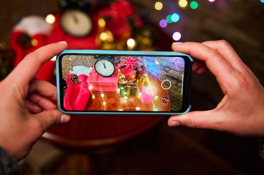 Mobile phone in live view. Female hands hold a smartphone, photograph a cozy Christmas interior with a black alarm clock with midnight on the dial, luminous garlands, lit candles and gifts on table