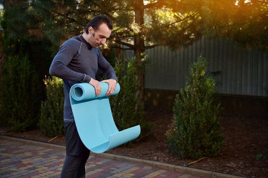 Active middle-aged Caucasian man rolling out the fitness mat. Beautiful sunbeams falling on the backyard. Male athlete exercising outdoor at sunset. Sport. Yoga practice. Active and healthy lifestyle