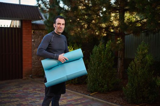 Handsome active Caucasian male athlete, sportsman, fit guy in his 40s holding a fitness mat, working out outdoors at sunset, smiling looking at camera, standing in the house backyard
