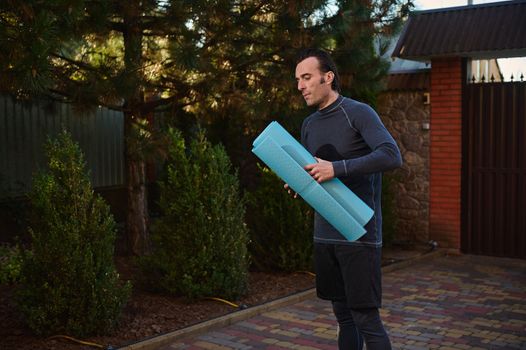 Portrait of a Caucasian man, a determined athlete, sportsman with a fitness mat while exercising outdoors, practicing yoga at sunset. Sport, active and healthy lifestyle.