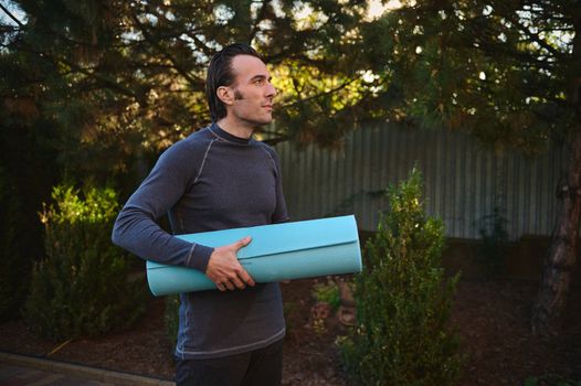 Side portrait of a Caucasian handsome active man in sportswear, posing with a blue fitness mat on the backyard, while practicing yoga at sunset. Attractive muscular male athlete working out. Sport