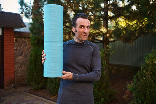 Portrait of an active Caucasian man, a determined athlete, sportsman with a fitness mat, smiles looking at the camera while exercising outdoors, practicing yoga at sunset. Sport and healthy lifestyle.