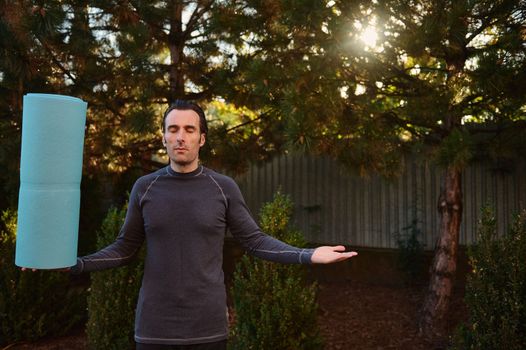 Peaceful calm Caucasian athlete man, yogi, sportsman holding a fitness mat, standing palms up in the backyard against a sunbeam. Spiritual growth. Mindfulness. Enlightenment. Yoga. Active lifestyle