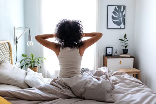 Rear view of unrecognizable young woman with curly hair waking up stretching sitting on bed ledge in cozy bedroom early in the morning. Lifestyle concept.