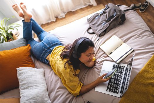 Top view of teenage asian college girl lying on bed using phone listening to music, podcast with wireless headphones. Doing homework, studying at home using laptop. Lifestyle concept.