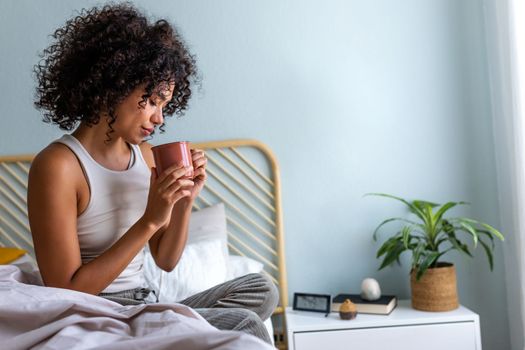 Young African American woman having morning coffee in bed. Multiracial latina female relaxing in bedroom having hot tea. Copy space. Lifestyle concept.