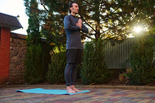Full length portrait of a handsome Caucasian sporty guy in active wear, exercising at sunset, meditating, standing barefoot on a fitness mat, holding hands at chest level, breathing deeply fresh air