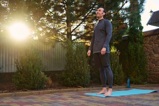 Full length active male athlete, sportsman, a yogi standing barefoot on a blue mat while practicing yoga outdoors at sunset. Copy advertising space. Active healthy lifestyle. Fitness. Sport. Workout