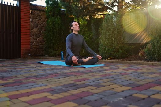 Peaceful sportsman, confident man yogi, athlete meditating in lotus pose outdoors, practicing yoga at sunset, while sun rays fall on backyard. Body care. Mindfulness. Enlightenment. Awakening