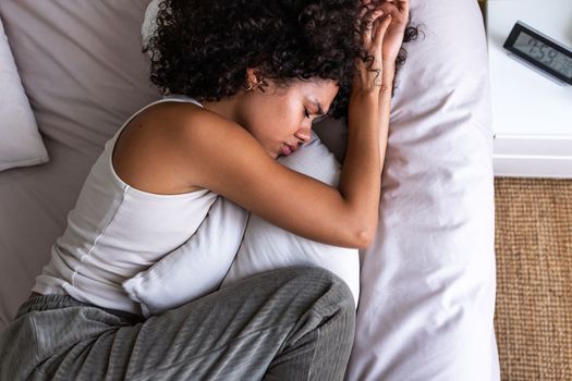 Top view of depressed and sad young African American woman lying down on the bed crying while hugging pillow. Sadness and mental health concepts.