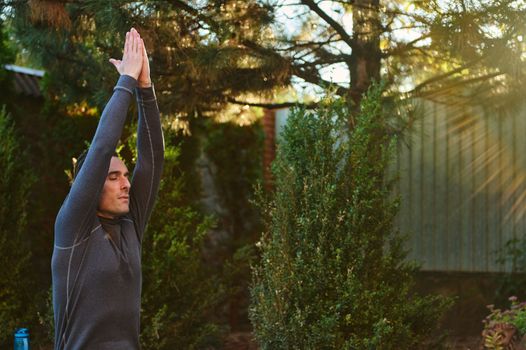 Active middle-aged Caucasian sportsman, male athlete, yogi raising hands to the sky, palm to palm, meditating, stretching his body, doing yoga at sunset, outdoors against coniferous trees background