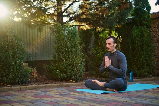 Sunset sunlight fall on the backyard, while a handsome active man, yogi meditating in lotus position, on a yoga mat, keeping palms together. Peaceful guy in gray sportswear breathing deeply fresh air.