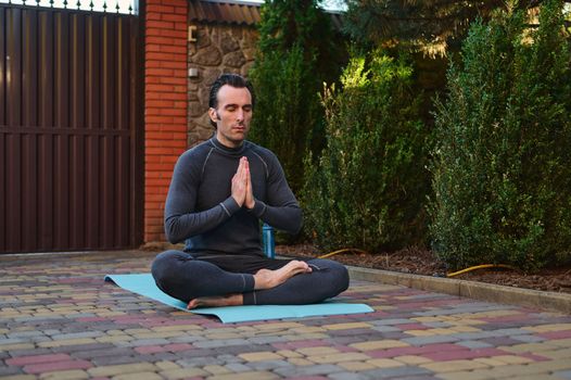 Peaceful Caucasian man athlete, a yogi meditating with his eyes closed in lotus pose, keeping hands palms together, sitting barefoot on a fitness mat. Yoga practice. Spiritual growth. Mindfulness