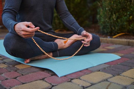 Cropped view of a sportsman, athletic man in gray sportswear, a yogi sitting in lotus pose on a blue fitness mat, holding holy rosary while meditating, practicing yoga outdoors at sunset