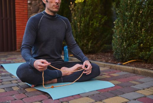 Cropped view of a yogi sitting on a fitness mat, meditating with rosary beads while practicing yoga outdoors. Body conscious. Sport. Active and healthy lifestyle. Meditation. Spiritual growth