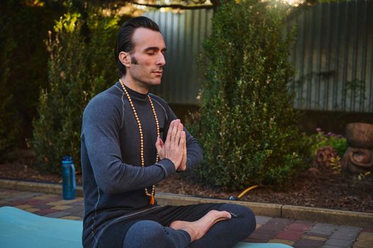 Peaceful man yogi sitting in lotus pose on fitness mat, keeping hands palms together while meditating with rosary beads outdoors at sunset. Yoga practice. Meditation. Body conscious. Healthy lifestyle