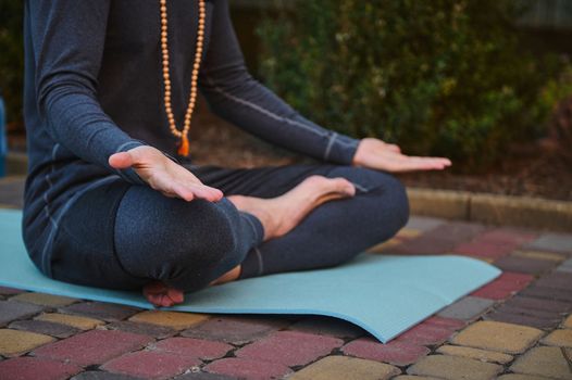 Cropped view of male athlete, yogi wearing rosary beads and gray sportswear, sitting on fitness mat in lotus position and palms up on knees, practicing yoga. Prayer, gratitude. Yoga pose. Meditation.