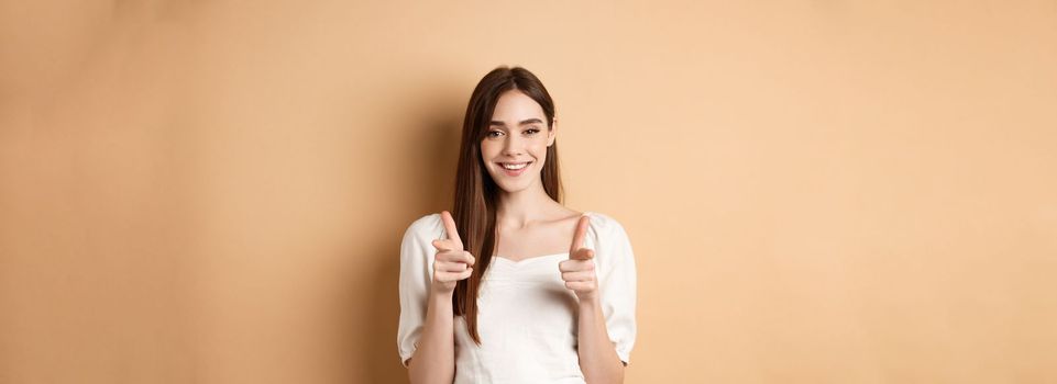 We need you. Smiling young woman pointing fingers at camera to beckon or invite people, standing on beige background.