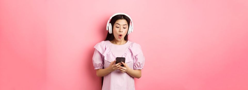 Excited asian girl looking at smartphone screen amused, say wow, listening music in wireless headphones, standing against pink background.