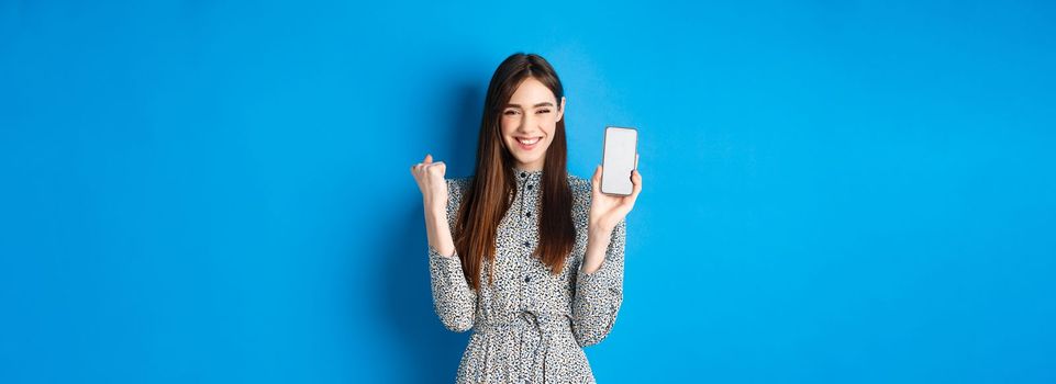 Cheerful pretty girl chanting, winning on mobile phone, showing empty smartphone screen and fist pump, smiling and celebrating, blue background.