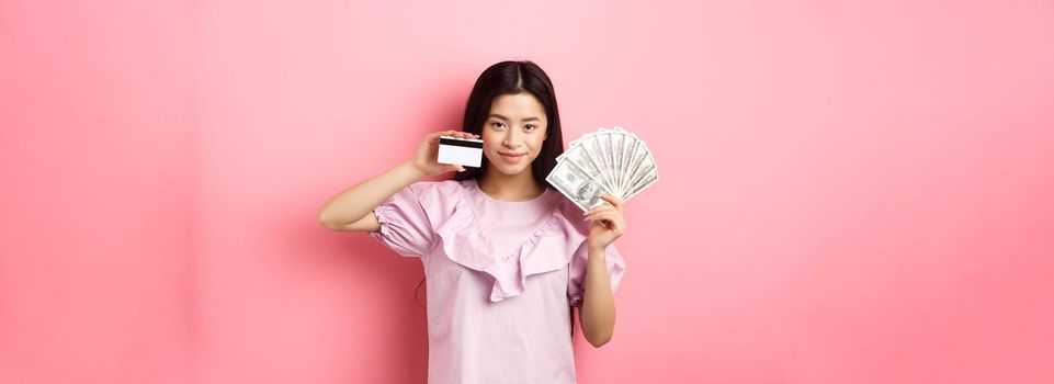 Smiling beautiful asian girl showing money and plastic credit card, giving choice, standing against pink background.