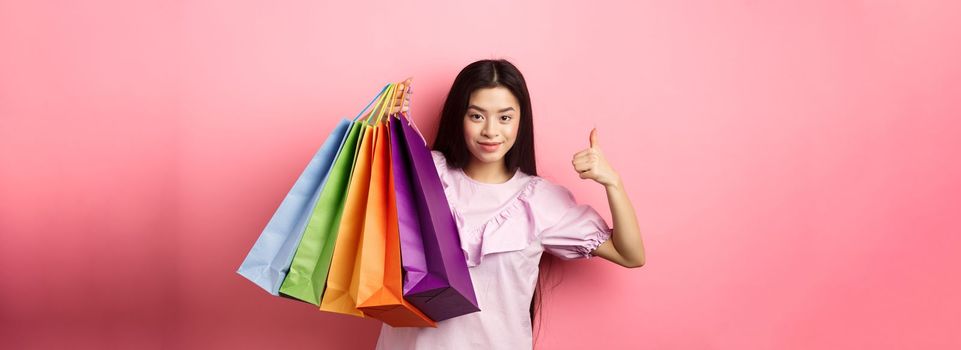 Shopping. Stylish girl showing colorful paper bags and thumbs-up, recommend store discounts, standing against pink background.