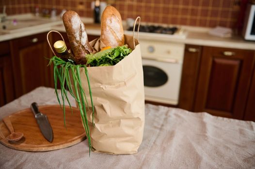 Delivered groceries, greens and loaves of wholesome bread on an eco shopping bag next to a wooden board on a kitchen table