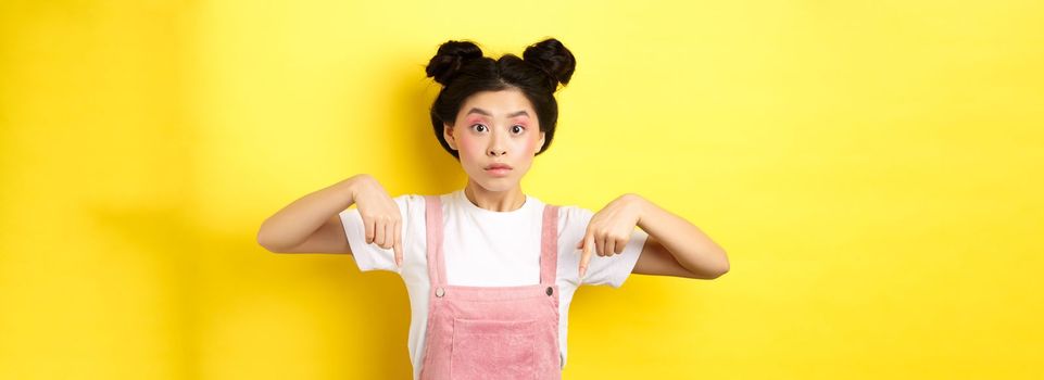 Surprised asian woman with beauty makeup, pointing fingers down and looking excited, showing interesting news, yellow background.