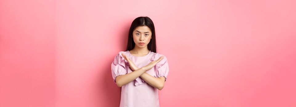 Serious chinese girl show cross sign, make stop gesture and look confident, block person, say no and reject offer, standing on pink background.