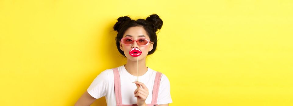 Summer and fashion concept. Stylish asian glam girl in sunglasses, holding party lips mask, standing on yellow background.