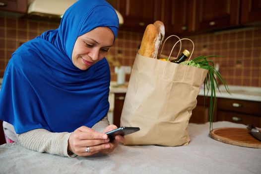 Beautiful Arab Muslim woman in hijab, using smartphone, shopping online, leaning against a kitchen table with a grocery shopping bag with healthy food. Healthy purchasing from grocery. Food delivery.