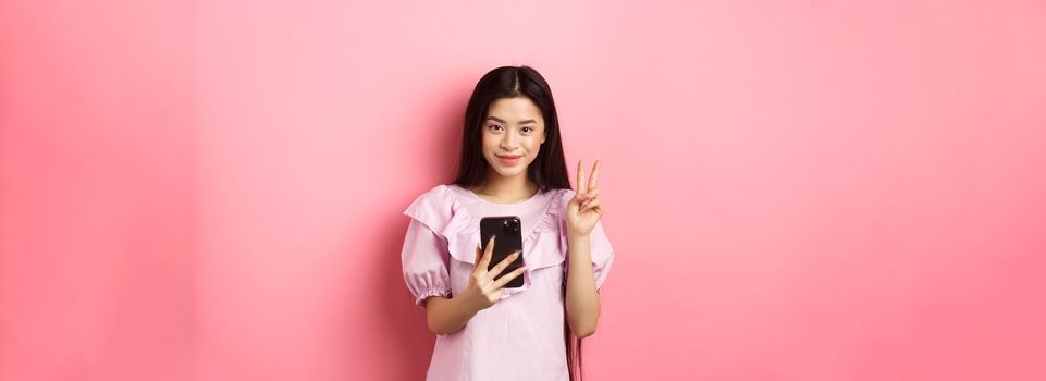 Online shopping. Stylish asian teen girl using smartphone, show peace sign and smiling pleased at camera, standing on pink background.