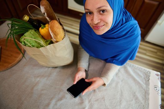 View from above of a pretty Middle-Eastern Muslim woman housewife in hijab, looks up at camera, checks expenses, using smartphone with copy advertising space for insert promotional text or mobile app