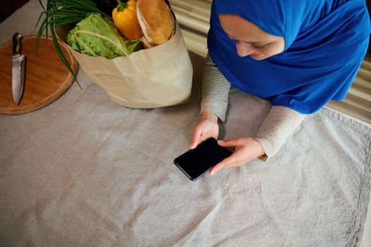 Top view woman housewife in hijab, arrives home with eco shopping bag with groceries and healthy food, checks expenses, using smartphone with copy ad space for insert promotional text or mobile app