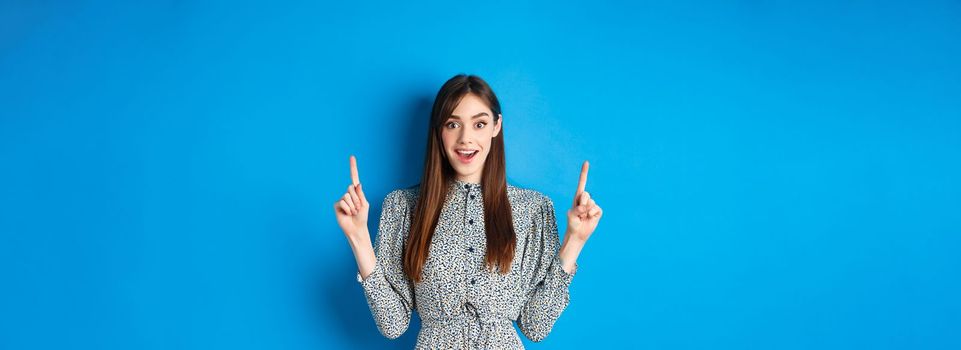 Excited and happy young woman in dress checking out special offer, pointing fingers up and smiling, standing against blue background.