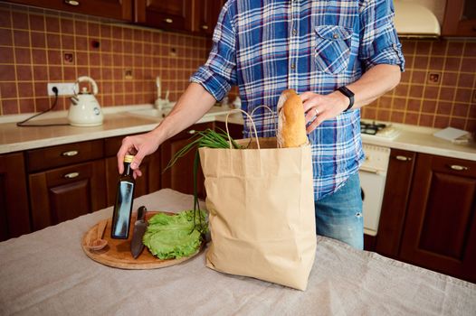 Cropped view Caucasian sporty man in blue checkered shirt, arriving home after shopping for grocery, standing at a kitchen island and unpacking groceries, sorting organic vegetables and healthy food