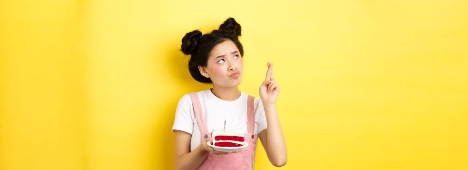 Holidays and celebration. Cute asian birthday girl making wish at party wish cake and candle, cross fingers and looking up, standing on yellow background.