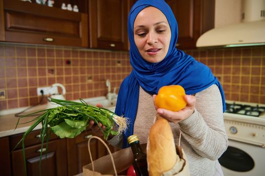 Happy young Muslim Middle-Eastern housewife in a blue hijab, lays out purchases in her cozy wooden home kitchen. Beautiful woman in good mood is sorting vegetables and fresh greens. Food purchasing