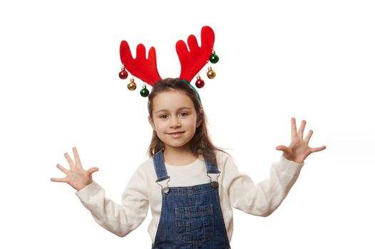 Isolated portrait on white background of a delightful Caucasian baby girl wearing white pull, blue denim overalls and deer antler hoop, expressing joy and happiness, cutely smiling looking at camera