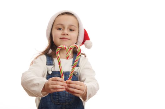 Selective focus on green and red stripped lollipops - candy canes in the hands of a blurred child girl wearing Santa hat and blue denim overrals, cutely smiling at camera, isolated on white background