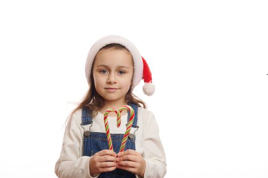 Lovely blue-eyed European little child girl in santa hat, holds striped christmas lollipops in the shape of heart, looking at camera isolated on white background. Copy space for christmas advertising