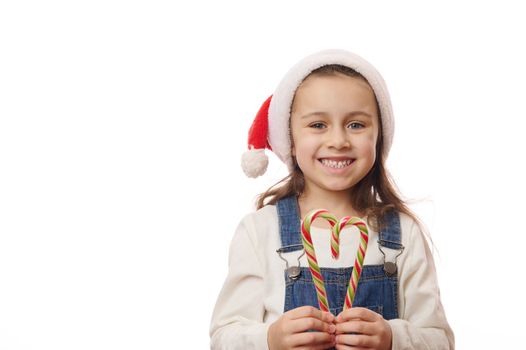 Lovely child, Caucasian baby girl in Santa hat, smiling a cheerful toothy smile at camera, posing with red and green striped candy canes over white background. Merry Christmas and New Year's concept