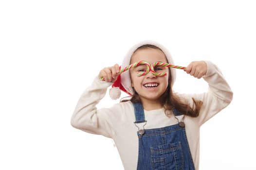 Mischievous baby girl in santa hat, smiling looking at camera through stripped lollipops like eyeglasses, isolated on white background. Merry Christmas and Happy New Year concept. Advertising space