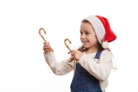 Caucasian 5-6 years child, cute baby girl in Santa hat, pointing with green red striped candy canes at copy advertising space for Christmas, on white background, expressing happiness and cheerfulness